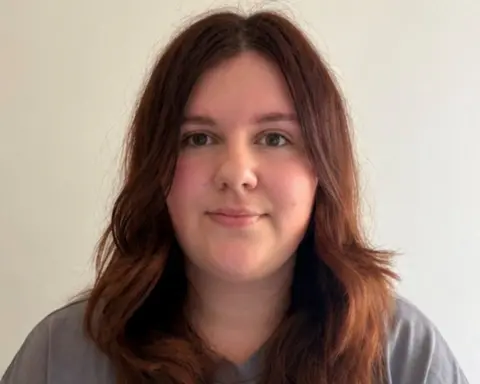 Faye Goodwin Faye, a woman with long brunette hair in a grey T-shirt, looks at the camera, standing in front of a plain white background