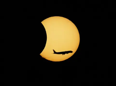 EPA/Shutterstock A plane flies in front of the Moon passing the Sun, which is a large yellow circle against the black sky 