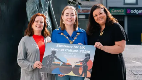 DCSDC Derry City and Strabane District Council Mayor Grace Uí Niallais is wearing a blue shirt and her mayoral chain. Deirdre Harte is wearing a red shirt and grey cardigan, while Jacqueline Whoriskey is wearing a long black dress. They are all holding the new branding for the Strabane Town of Culture bid.