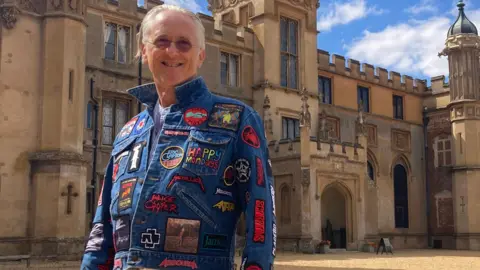 Justin Dealey/BBC A man with grey hair is stood outside Knebworth house wearing a blue denim jacket covered in band badges