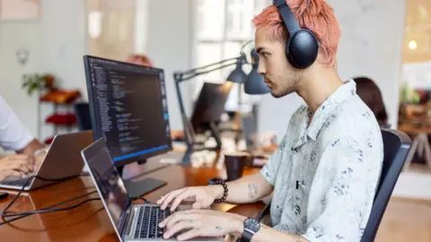 Getty Images A tech worker with pink hair sits at a desk and works on a laptop