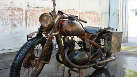 German War Graves Commission A rusted but largely well-preserved World War Two-era Wehrmacht motorcycle pictured parked in a building resembling a warehouse. 