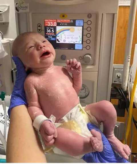 Maisie Savage Newborn baby in the foreground, wearing only a disposable nappy and supported by gloved hands in front of a neonatal warming unit. A hospital identification band is attached to the baby’s wrist. The warming unit’s display screen shows temperature settings and a weight reading, with control buttons and gauges visible around it. Medical equipment, tubing and wall-mounted hospital fittings appear in the background. The image is taken in a clinical hospital setting.