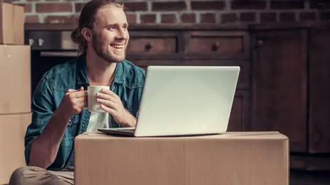 Getty Images Smiling man sits on the floor surrounded by boxes, with a laptop on a box in front of him and a cup of tea in his hand.