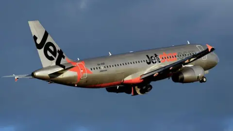 Getty Images A silver plane with Jetstar's orange and black livery flies against a dark cloudy sky.