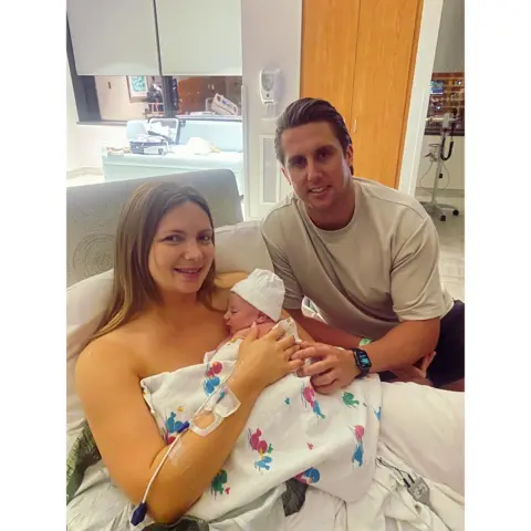 Maisie Savage Newborn baby in the foreground, wearing a white knit hat and wrapped in a white hospital blanket with colourful cartoon prints, held against the chest of a person sitting upright in a hospital bed. An IV cannula and tubing are attached to the person’s forearm. Another person in a light-coloured short-sleeved top sits beside the bed with one hand resting on the blanket and a smartwatch on their wrist. White bedding, pillows and a hospital room with cabinets, a sofa, medical equipment and windows are visible in the background.