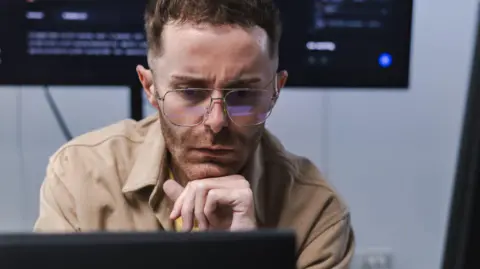 Getty Images A software engineer, wearing a light brown shirt, looks at a computer monitor in front of him with a concerned expression. A larger screen with computer code on it is on a wall behind him.