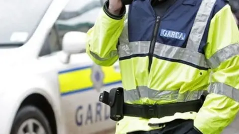 BBC Irish police officer, standing in a high-vis top with Garda written on it. A white Garda car is on the left.