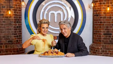 BBC A woman wearing a yellow dress, with her long blonde hair styled half-up, and a man with grey hair, wearing a black blazer and black shirt, sit in front of a brick wall with the MasterChef logo in the centre, an M surrounded by black, white, yellow and blue circles, eating a large plate of spaghetti and meatballs.