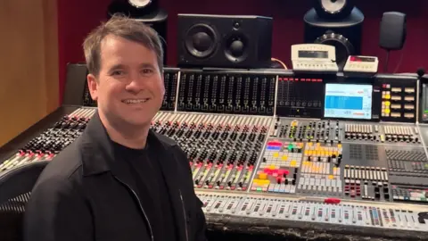Supplied Chris Peabody with short brown hair, a black top and black coat, seated at a large professional audio mixing console in a recording studio. The console is covered with rows of faders, knobs, buttons, and illuminated controls. Multiple studio monitor speakers are positioned around the workstation, and two small display screens sit above the mixing desk. The room features dark red walls and warm lighting, highlighting the extensive audio production equipment.