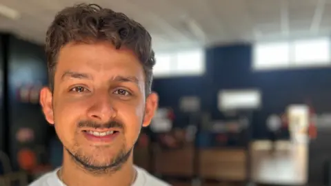 Chris is a 30 year old man with a dark beard and dark hair that is curly on top. He is wearing a white t-shirt with a silver chain necklace and is standing on a wooden dance floor.