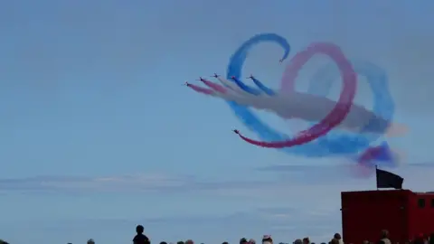 Aeroplanes doing a loop create a heart shape in the sky with blue and red jet smoke. Crowds watch below. 