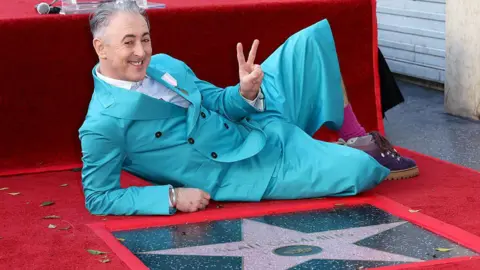 Getty Images Alan Cumming, wearing a turquoise suit, poses on the ground behind his Hollywood Walk of Fame star. He is on one side, supported by one elbow, with the other hand raised in a two-finger peace sign. 