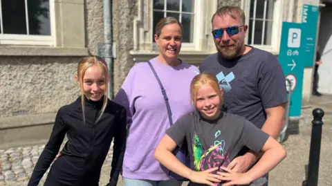 BBC A family of four smile at the camera, the two girls have blonde hair, the one on the left is wearing a black top, the one on the right is wearing a grey top with blue yellow and pink design. Behind the woman is wearing a purple jumpers and man is wearing a navy top and sunglasses.