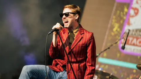 Getty Images Ricky Wilson of the Kaiser Chiefs, dressed in jeans and a yellow striped red blazer, wearing a yellow shirt and sunglasses, singing into a leaning microphone stand.