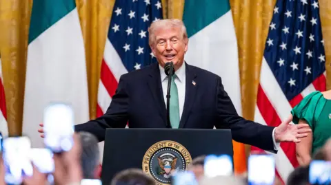 EPA/Jim Lo Donald Trump wearing dark suit and green tie. Standing at a podium in front of Irish and American flags. Trump has grey hair.