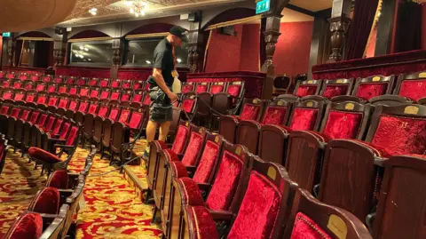 BBC/Richard Moss A man in black t-shirt and shorts and a baseball cap is cleaning carpets in the Theatre Royal with a hose and bottle. The carpets have a gold and red pattern and the seats are red and plush.