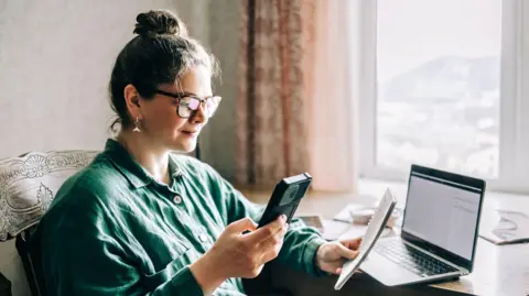 Getty Images Woman sits at a window seat holding a phone and a pad with a computer on a table in front of her.