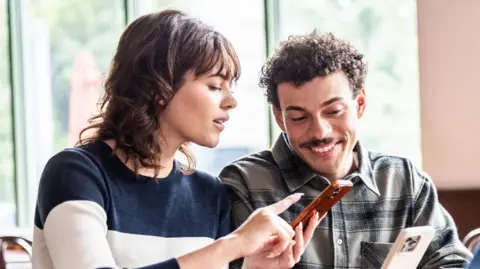 Getty Images A woman with dark shoulder-length hair points at a smartphone held by a smiling man with curly hair and a moustache.