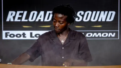 Luke Fowler Hugues Sio stands behind the decks while DJing. He is wearing a black short-sleeved shirt, headphones and a silver necklace. Behind him is a black and white sign which says 'RELOAD SOUND Footlocker v Solomon'