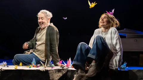 Tommy Ga-Ken Wan Two performers sit on the edge of a stage set, surrounded by brightly coloured origami cranes scattered around and suspended in the air. Dramatic spotlights illuminate the scene against a dark background, drawing attention to the vivid paper sculptures and casual costumes.
 The theatrical setting creates a striking contrast between the simple staging and the whimsical, dreamlike display of floating paper birds.
