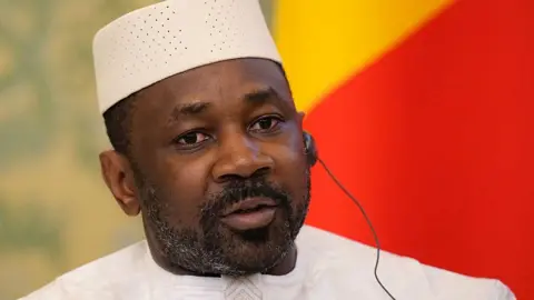 AFP via Getty Images Mali's junta leader attends a meeting with Russia's President at the Kremlin in Moscow. He wears a white outfit and communicated with the help of a translation earphone. Behind him is the national flag of Mali.