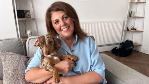 BBC Glynda Cullen with her pet Dachshund, Lemon, sit on a sofa in her living room. Glynda has brown hair and wears a blue blouse. 