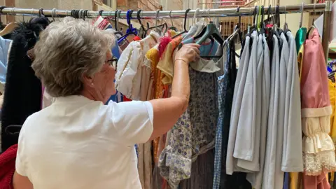 Alex Pope/BBC A woman going through a rack of clothes. She is holding onto a hangar and is wearing a white shirt, has short hair, is wearing glasses and has her back to the camera.