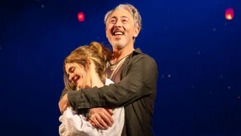 Tommy Ga-Ken Wan Two performers stand together on a dimly lit stage, framed by a deep blue backdrop and glowing red lanterns suspended overhead.
Warm spotlighting highlights the embrace, while the floating lights create a dreamlike, theatrical atmosphere.