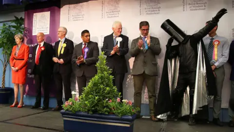 Getty Images A row of candidates can be seen on stage at the election count. Theresa May is on the far left of at least nine candidates on stage. She wears a bright red skirt suit and matching high heels, with her grey hair in a neat bob. She has her hands clasped on front of her and is smiling wryly. Lord Buckethead is on the far left of the row of candidates and is doing a dab. He wears a large black bucket on his head, with black armour and a black cape with a sliver lining.