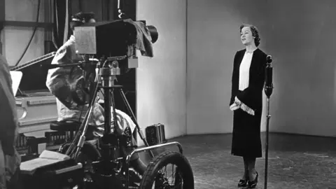 BBC A black and white image from 1936 - a smartly dressed lady stands in front a microphone and a television camera 