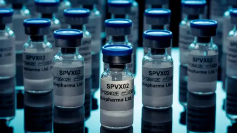 StablePharma This image shows multiple small glass pharmaceutical vials arranged on a reflective surface under cool blue lighting. The vials have blue caps and white labels, with the text “SPVX02” clearly visible on several containers; additional smaller text appears to include a product code and the company name Stablepharma Ltd. The composition places one vial in sharp focus near the center foreground, while numerous similar vials extend into the background with progressively softer focus. The vials appear to contain a white powder. Reflections of the vials are visible on the glossy surface beneath them, creating a symmetrical, laboratory-style aesthetic. The color palette is dominated by blues, whites, and blacks, and the lighting emphasizes the glass, metal, and plastic textures of the containers. The overall setting resembles a pharmaceutical, biotechnology, or medical research environment, with the image focusing on the repeated pattern of labeled drug or vaccine vials.
