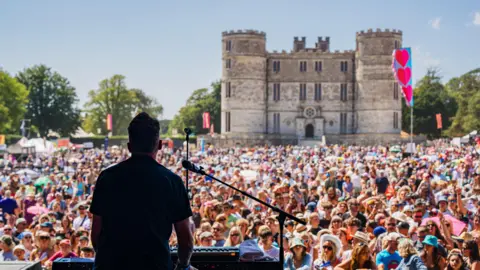 Camp Bestival A performer is on stage at a keyboard and microphone with his backturned. Beyond him is an enormous crowd of people. Behind the crowd there is a large castle. 
