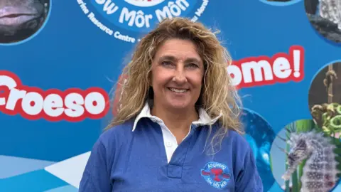 Frankie Hobro, a woman in her early 50s with blonde hair, looking at the camera.  She's wearing a blue polo shirt with the logo of the Anglesey Sea Zoo and standing in front of an Anglesey Sea Zoo banner