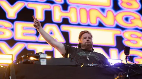 Getty DJ Eats Everything performing on the stage. He is standing behind the decks and has one arm raised in the air. The screen behind him says 'EATS EVERYTHING' in yellow and pink writing.