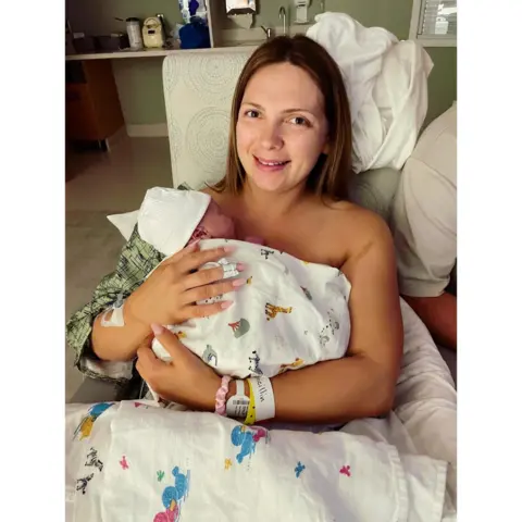 Maisie Savage Newborn baby wrapped in a white patterned hospital blanket and wearing a white knit hat, held against the chest of a person seated in a hospital bed. The person has a hospital wristband and an adhesive dressing on one wrist. White bedding and pillows surround the bed. A partially visible person in a white shirt sits beside them. In the background, a hospital room contains a sink, countertop and storage area with medical and personal items.