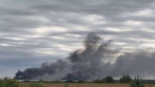 Dark smoke rises above a horizon of dry grass. The sky is grey.