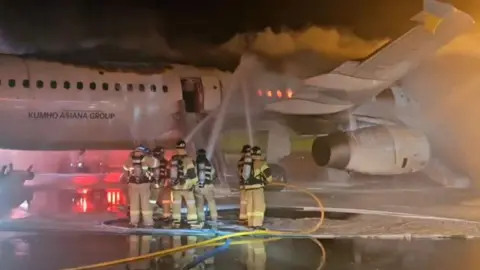 Yonhap via Reuters Six firefighters train hoses on the burning fuselage of a Air Busan plane at Gimhae Airport, South Korea.