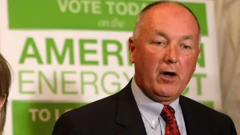 Getty Images Pete Hoekstra, with short grey hair, and in a dark suit coat, red tie and patterned blue shirt, stands in from of a green and white campaign sign promoting American energy. 