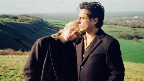 Amber Little An image of a blonde woman who is wearing all black leaning on a man with brown hair who is also wearing all black in the middle of a field.