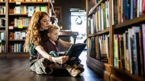 Getty Images A stock image of a woman with curly hair reading a story to a young boy sitting in her lap. They are in a library. 