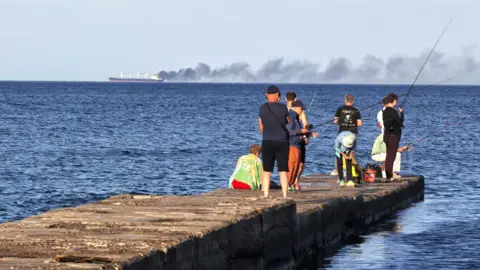 Getty Images Men fish on a pier as black smoke rises from a vessel damaged by a Russian attack in the Black Sea, Odesa region, Ukraine, July 28, 2026