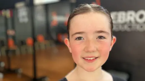 BBC Ten year old Martha is looking at the camera, she is fair skinned with freckles and her hair is in a tight bun. Behind her is a dance studio. 