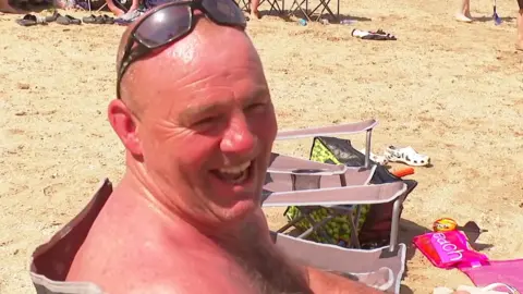 A sunburnt man sits in a chair on the beach. He is bald, topless and has sunglasses on his head. 