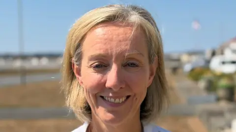 BBC A woman called Ruth Thomason smiles at the camera while standing outside an office in Guernsey. She has fair hair and the top row of her teeth are showing.