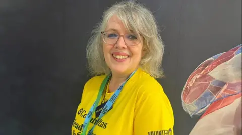 Volunteer now A woman, Camilla Reynolds pictured In front of a black wall with blue, white and red wall art on the right corner.  She has grey hair and is wearing a yellow t-shirt with a blue lanyard and is wearing red glasses. 