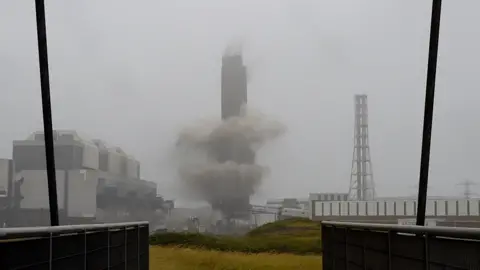 Smoke surrounds the a tall chimney as it is demolished. It is a grey, foggy day with a white sky behind the chimney. A building can be seen to its left-hand side. 