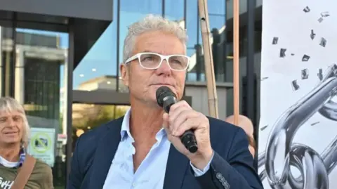 EPA A man wearing white-rimmed glasses and a dark suit, holds a microphone