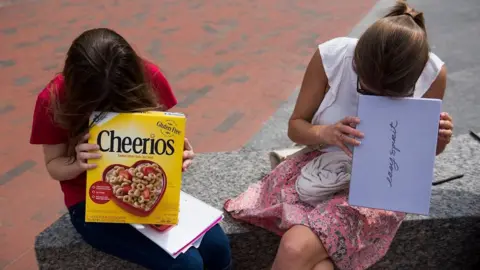 The Boston Globe/Getty Images Two girls look into cardboard boxes, one a cereal box and another a shoe box, while sitting on a bench outside.