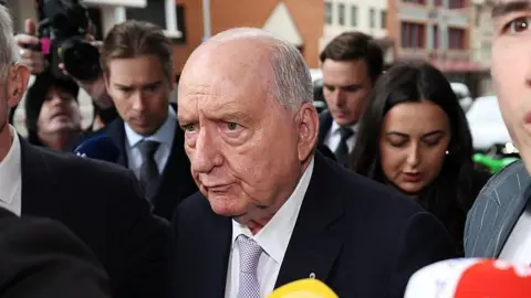 Getty Images An old man, partially bald with short grey hair, wearing a dark suit and surrounded by a crowd